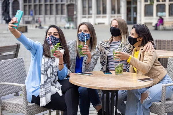 Antwerp, Belgium, May 21, 2021, Multiracial young female friends wearing face mask while taking selfie with mobile smartphone cam while drinking cocktails on a terrace of a cafe in the city during — Stock Photo, Image