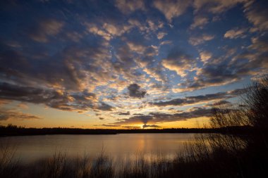 Önünde ağaç siluetleri olan güzel mavi bir göl dramatik bir günbatımı gökyüzünün altında.