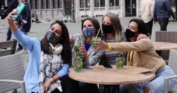 Antwerpen, Belgien, 21 maj 2021, Multiraciala unga kvinnliga vänner bär ansiktsmask samtidigt som de tar selfie med mobil smartphone kamera medan du dricker cocktails på en terrass i ett café i staden under — Stockvideo