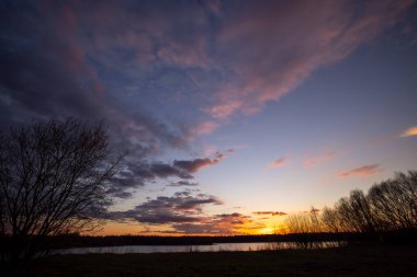 Önünde ağaç siluetleri olan güzel mavi bir göl dramatik bir günbatımı gökyüzünün altında.