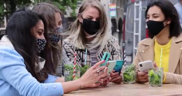 Junge Freundinnen verschiedener Rassen tragen Gesichtsmasken per Handy, während sie während des Coronavirus-Ausbruchs auf der Terrasse eines Cafés in der Stadt einen Drink trinken - Ernährungs- und Technologiekonzept — Stockvideo