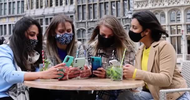 Jóvenes amigas multiraciales que usan mascarillas con smartphone móvil mientras toman una copa en la terraza de un café de la ciudad durante el brote del virus corona - Concepto de comida y tecnología — Vídeos de Stock