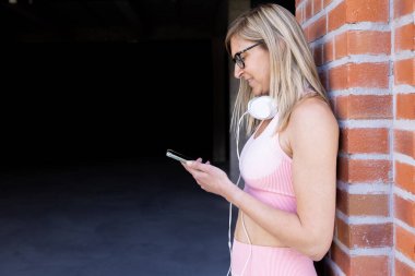 Egzersiz yaptıktan sonra dinlenirken akıllı telefon ve kulaklık kullanan, dışarıda duvara yaslanan genç ve güzel bir kadın. Teknoloji, egzersiz konsepti