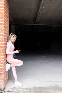 Egzersiz yaptıktan sonra dinlenirken akıllı telefon ve kulaklık kullanan, dışarıda duvara yaslanan genç ve güzel bir kadın. Teknoloji, egzersiz konsepti