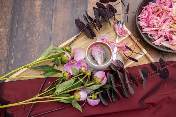 Белая чашка черного кофе стоит на деревянном фоне рядом с розовыми пионами и другими садовыми цветами и листьями, концепция деревенской жизни, Cottagecore, ruralcore, cluttercore. — стоковое фото