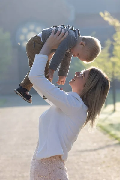 Молодая счастливая мать с маленьким ребенком на руках, глядя на него с украшением и любовью, стоит посреди парка под дождем восходящего утреннего солнца — стоковое фото
