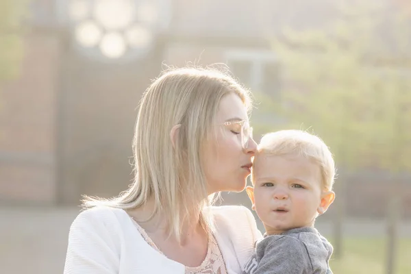 Молодая счастливая мать с маленьким ребенком на руках, нежно целуя его голову, стоит посреди парка в лучах восходящего утреннего солнца — стоковое фото