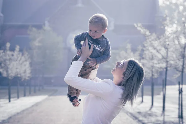 Молодая счастливая мать с маленьким ребенком на руках, глядя на него с украшением и любовью, стоит посреди парка под дождем восходящего утреннего солнца — стоковое фото