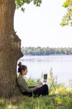 Herhangi bir yerden çalış. Uzaktan kumandalı doğada çalışıyor. Genç bir kadın, dizüstü bilgisayarla çalışan serbest çalışan güzel bir orman ve göl manzaralı bir kadın. Bağımsız cennet, tele-çalışma özgürlüğü. uzaklık