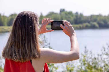 Kırmızı etekli güzel beyaz kadın dışarıdaki yeşil orman gölünde cep telefonuyla işaret fişeğiyle fotoğraf çekiyor. Yüksek kalite fotoğraf