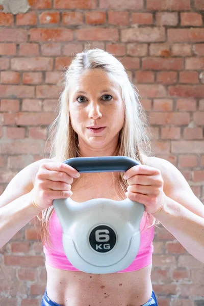 Blonde young muscular woman with sporty looks doing high pull exercise with kettlebells on hard training at the garage gym. High quality photo