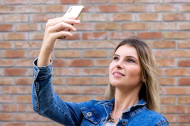 Neşeli, sevimli, sevimli, güzel, çekici kız arkadaş sarışın, soluk tenli, elinde telefonuyla selfie çeken kırmızı renkli tuğla üzerine izole edilmiş kot ceket giyen.