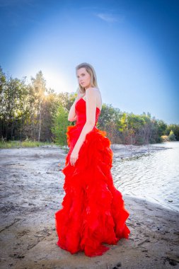 Beautiful and sensual young woman wearing a red evening dress outside in a natural setting against a mountains and lake background. High quality photo