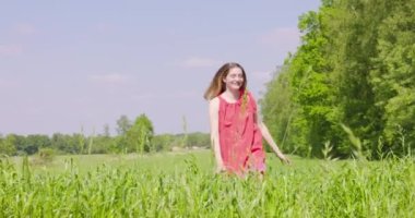 A young girl happily walking through a field touching with hand wheat ears. Beautiful carefree woman enjoying nature and sunlight in wheat field at incredible colorful sunset. High quality 4k footage
