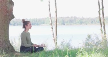 Yoga yapan genç bir kadın. Bir ağaç ve bir orman gölünün yanındaki yeşil çimlerin üzerinde oturuyor. Egzersiz yapmak ve sağlığınızı korumak için iletişim kurun. Yüksek kalite fotoğraf