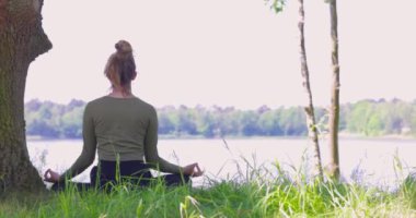 Yoga yapan genç bir kadın. Bir ağaç ve bir orman gölünün yanındaki yeşil çimlerin üzerinde oturuyor. Egzersiz yapmak ve sağlığınızı korumak için iletişim kurun. Yüksek kalite fotoğraf