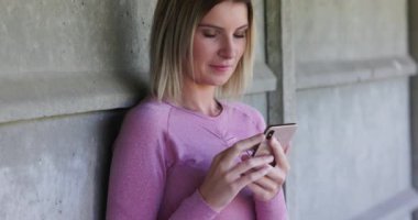 Egzersiz yaptıktan sonra dinlenirken akıllı telefon kullanan, dışarıda duvara yaslanan güzel bir kadın. Teknoloji, egzersiz konsepti. Yüksek kalite 4k görüntü