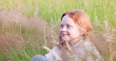 Yaz tarlasında çimlerin üzerinde oturan, güneşin tadını çıkaran ve saçlarını düzelten uzun kızıl saçlı tatlı bir genç kız. Yüksek kalite 4k görüntü