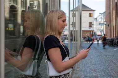 Mutlu sarışın milenyum kızı, modaya uygun giysiler içinde güneşli bir günde şehir ortamında gülen, Avrupa 'da sıradan görünümlü bir şehirde elinde akıllı telefon cihazıyla gezen neşeli bir kadın.