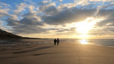 Bulutlarla ve altın güneşle boyanmış bir gökyüzünün altında, denizin kumlu sahilinde muhteşem bir gün batımında yürüyen iki kişinin siluetleri. Resimli doğa manzarası. Bulutlar yansıyor.