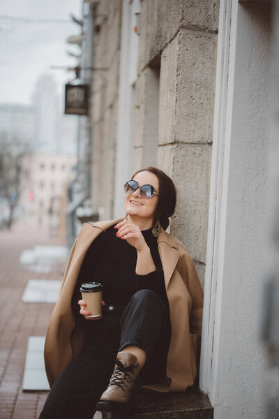 Portrait of a stylish woman dressed in a beige raincoat and sun goggles drinking a cup of coffee cloudy on the building. Street fashion concept.