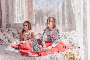 Küçük bir oğlan ve kız kanepede oturuyorlar. Kırmızı bir halı ve bir süs ile kaplılar. Çocuklar iletişim kurar, kanepede otururken güler. Oda güneş ışınlarıyla doludur. Erkek ve kız kardeş.