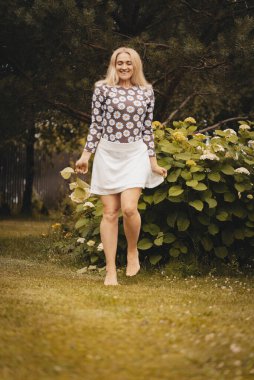 Beyaz etekli, sarı saçlı ve yaz süveteri giyen mutlu kadın parkta yalınayak yürüyor. Dışarıda çiçek tutan bir kız. Mutlu kız sıcak yaz havasının tadını çıkarıyor. Kız çimenlerde koşuyor..