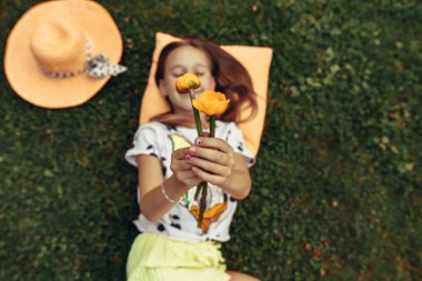 Yukarıdan bir fotoğraf, küçük bir kız elinde çiçeklerle yeşil çimlerin üzerinde yatıyor, hasır bir şapkanın yanında. Doğada vakit geçirmek. Parktaki açık havada bir çocuğun portresi. Seçmeli odak, ayrışma.