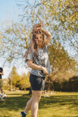 Sıcak bir yaz gününde, kalın kıvırcık saçlı, tişörtlü ve siyah şortlu mutlu genç kız parkta yürüyor. Güneşin parlak, sıcak ışınları. Yaz parkında yürüyüş. Gülümseyen kız açık havada.