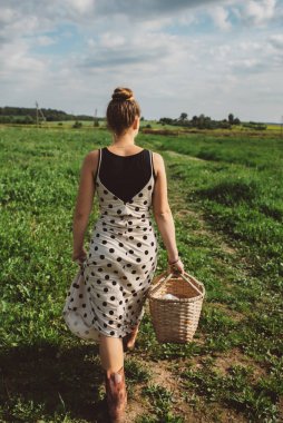 Uzun bir yaz elbisesi içinde elinde sepetle pikniğe giden genç bir kızın sırtından bir fotoğraf. Yaz tatili. Açık mavi gökyüzü. Defoktus, seçici odaklanma.