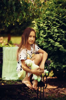 Bir yaz parkında elinde yumuşak bir oyuncakla bir bankta oturan etekli küçük bir kız. Doğada vakit geçirmek, yaz tatilleri. Parktaki açık havada bir çocuğun portresi. Seçmeli odak, ayrışma.