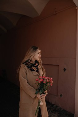 Elinde bir buket kırmızı gülle uzun sarı saçlı, gülümseyen genç bir kız şehirde dolaşıyor. Bahar yürüyüşü, güneşli bir gün. Elinde çiçekler olan bir kadın..