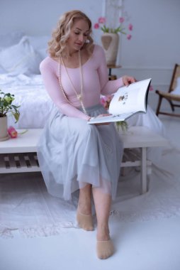 Uzun, soluk pembe etekli kısa kıvırcık saçlı tatlı bir kız parlak bir odada beyaz bir yatakta oturup dergi okuyor. Ev bitkileri. Kızın boynunda uzun inci boncuklar var..