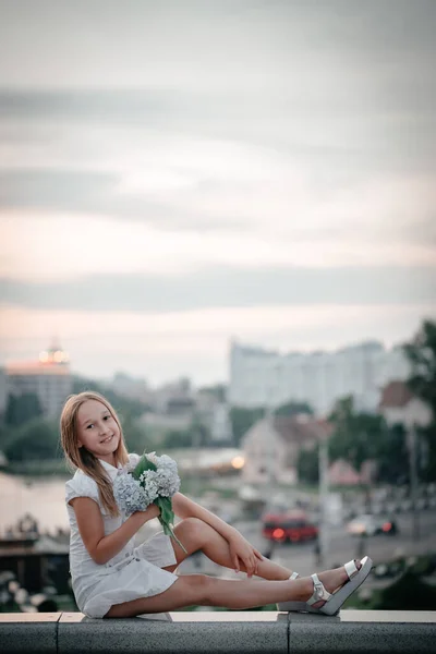 Dziewczynka Białej Sukience Kwiatami Rękach Siedząca Balustradzie Mostu Tle Miasta — Zdjęcie stockowe