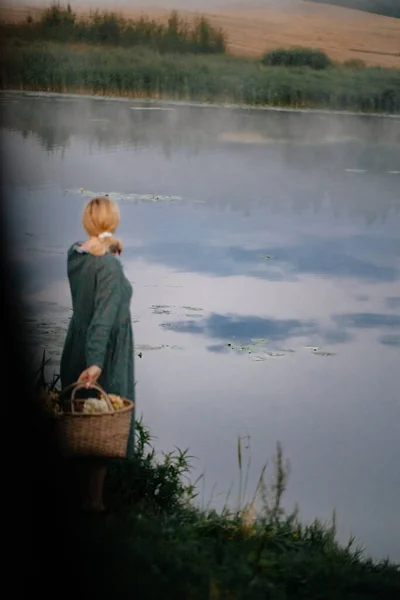 一个穿着长裙 手里拿着稻草篮的女孩正沿着一条安静的河边走着 一个女孩一大早在湖边散步 一筐花 有选择的重点 分散重点 — 图库照片