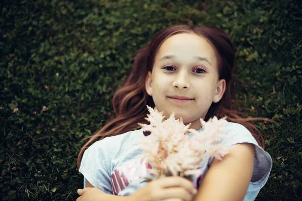 写真は 長い暗い髪の女の子の上から彼女の手の中に花が草の上に横たわってポーズをとった 屋外で花を持つ子供の肖像画 選択的フォーカス フォーカス トーン — ストック写真