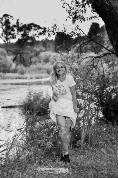 白いチュニックとスタイリッシュな裂けたジーンズのブロンドの髪の笑顔の女の子が湖の岸に立っています リラクゼーションと楽しみの概念 夏のカップル 湖のそばの森 — ストック写真