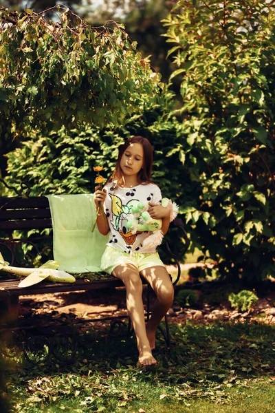 一个穿着裙子的小女孩坐在夏季公园的长椅上 手里拿着一个柔软的玩具 在大自然中度过的时光 公园里一个孩子的画像 有选择的重点 分散重点 — 图库照片