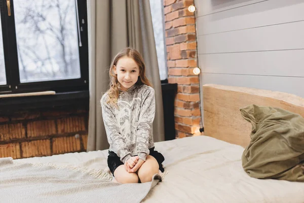 暗い巻き毛の少女が彼女の部屋のベッドの上に横たわっている 背景に黄色のガーランド 子供はベッドの上で寛いでいる 近代的な寝室のインテリア 照明で装飾された居心地の良い部屋 — ストック写真