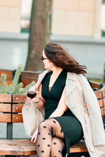 一个身穿黑色连衣裙和米色风衣的年轻漂亮的姑娘手里拿着杯酒在城里走来走去 街头时尚概念 — 图库照片