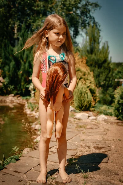一个穿着泳衣的小女孩在露天池塘边玩洋娃娃 阳光灿烂的一天 一个女孩在池塘里游泳 — 图库照片