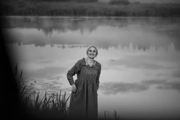 一个金发女郎穿着长裙沿着一条安静的河岸走着 女孩一大早在湖边散步 一个快乐女人在户外的画像 有选择的重点 分散重点 — 图库照片