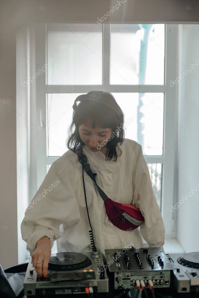 Portrait of a young stylish girl DJ in a white loose shirt and headphones, playing hop-hop on turntables. DJ makes music. DJ mixer. Photo with selective focus.
