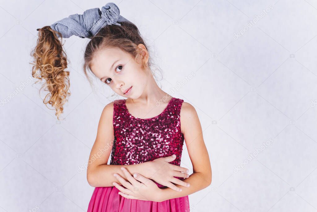 Cute little girl with long dark hair gathered in a ponytail in a pink dress with sequins posing on a white background. The child is isolated on a white background. A child with curly hair.