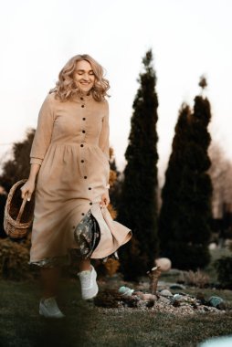 A sweet girl with blond curly hair in a long dress with a basket in her hands is walking in the park among the trees, spring has come. Walk in the park. Landscaping. Defocus.