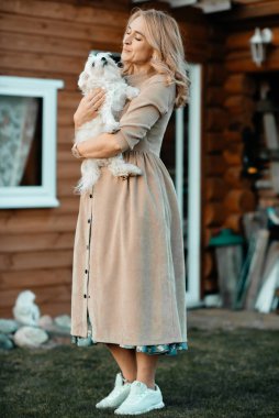 A blonde girl with curly hair in a long dress holds a small white dog in her hands while standing in the open air against the background of a wooden house. Pedigree dog. Pets concept. White puppy.