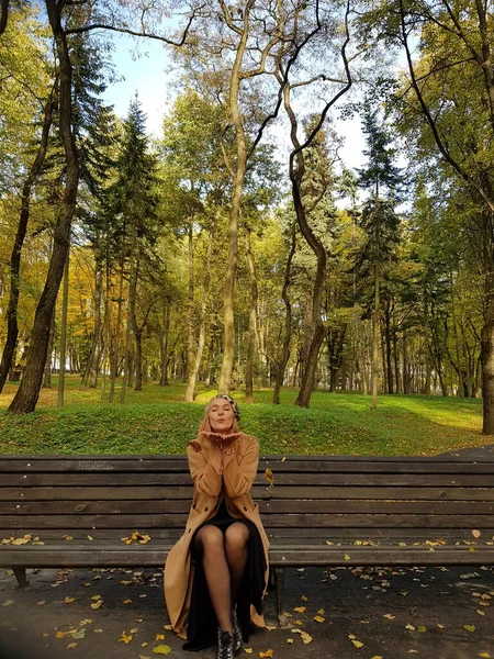 A stylish girl in a light coat with a beret on her head sits on a bench in an autumn parkra on a sunny day. Autumn time, yellow leaves on the asphalt. The lady is fanning the autumn foliage.