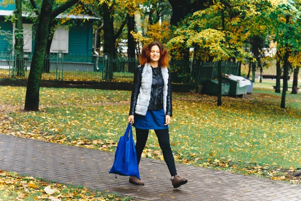 Pofuduk saçlı, elinde mavi bir resim olan mutlu bir kız sonbahar parkında yürüyor. Sonbahar yaprakları yere düştü..