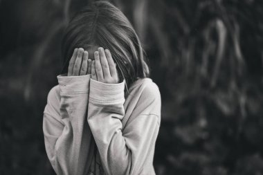 Yüzünü elleriyle örten gri kazaklı küçük bir kızın portresi. Korkmuş çocuk, korku. Seçmeli odak, ayrışma.