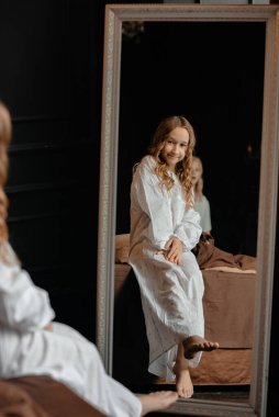 Beyaz pijamalı, siyah kıvırcık saçlı tatlı bir kız büyük bir aynayla yatakta oturuyor. Çocuk aynaya bakıyor. Seçici odaklanma, defoux.